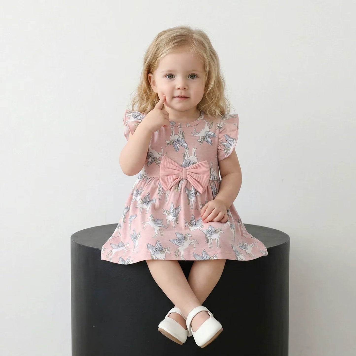 A toddler girl with blonde hair sits on a black cylindrical pedestal wearing a pink flutter-sleeve dress with a blue and white butterfly print and a large pink bow at the waist. She is wearing white Mary Jane shoes against a plain white background.