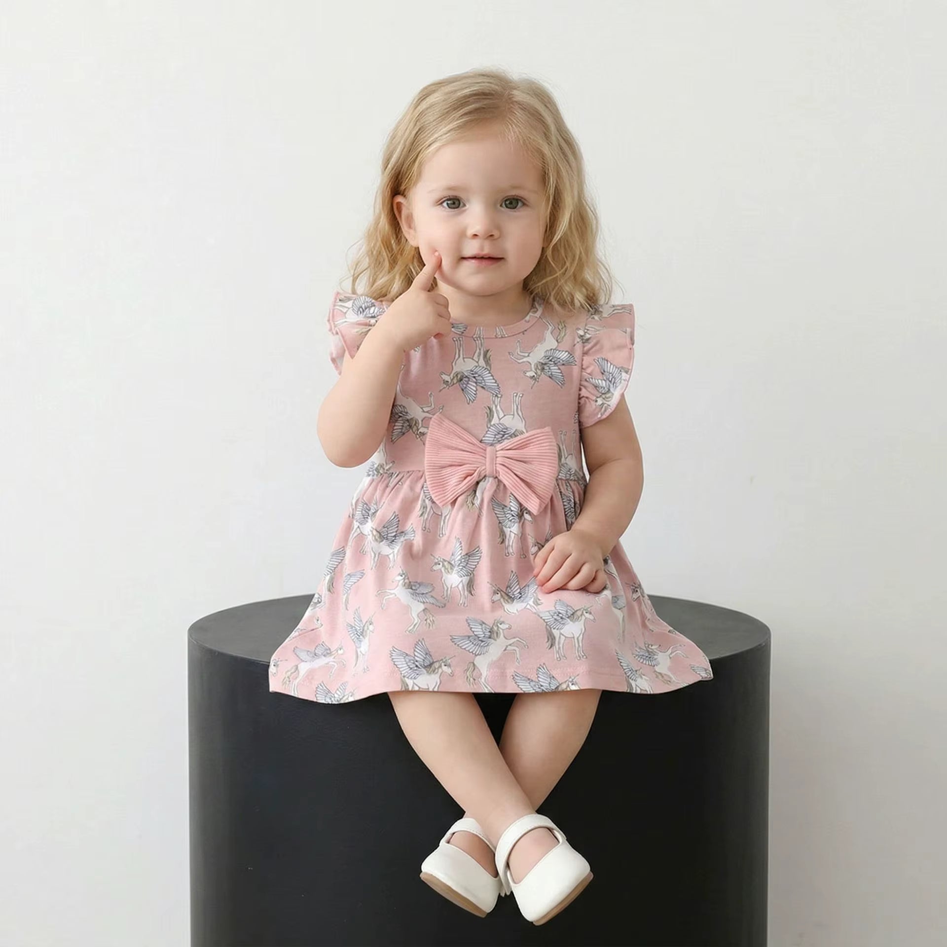 A toddler girl with blonde hair sits on a black cylindrical pedestal wearing a pink flutter-sleeve dress with a blue and white butterfly print and a large pink bow at the waist. She is wearing white Mary Jane shoes against a plain white background.