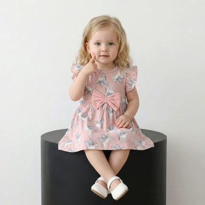 A toddler girl with blonde hair sits on a black cylindrical pedestal wearing a pink flutter-sleeve dress with a blue and white butterfly print and a large pink bow at the waist. She is wearing white Mary Jane shoes against a plain white background.